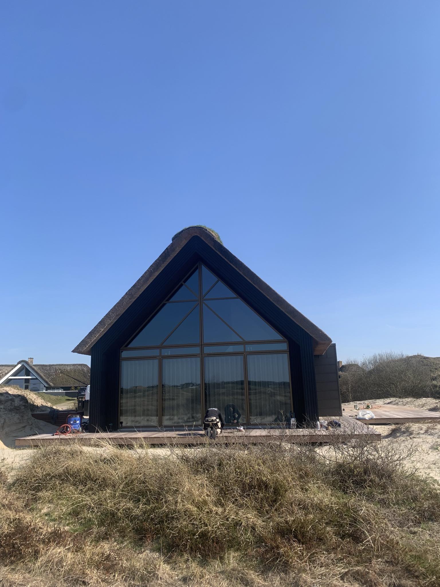 Sommerhus med sort beklædning, stråtag og glasgavl i klitterne på Fanø
