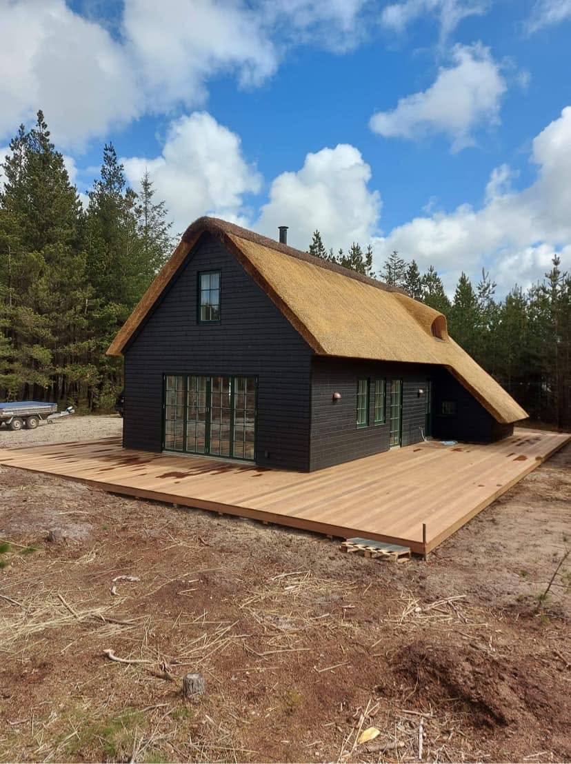Sort sommerhus med stråtag og terrasse mellem fyrretræer